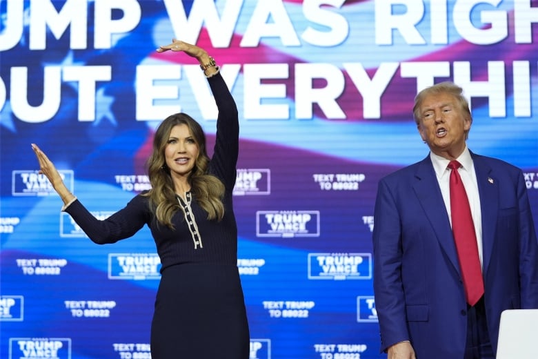 A woman makes a C-shape with her arms on a  stage beside a man in a  suit