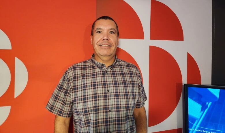 A man stands in front of a red and white background. He is wearing a grey plaid shirt.