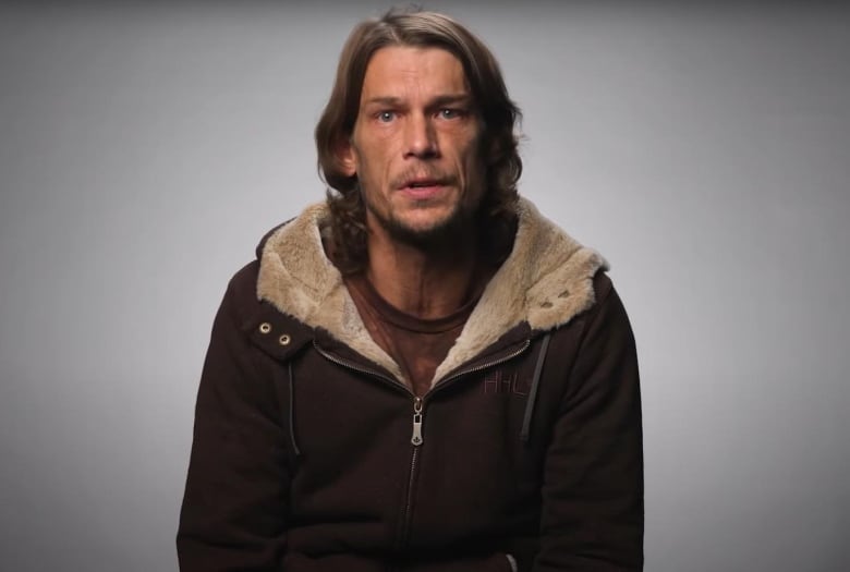 A man stands in front of a grey and white background. He is wearing a brown jacket over a light brown shirt. 