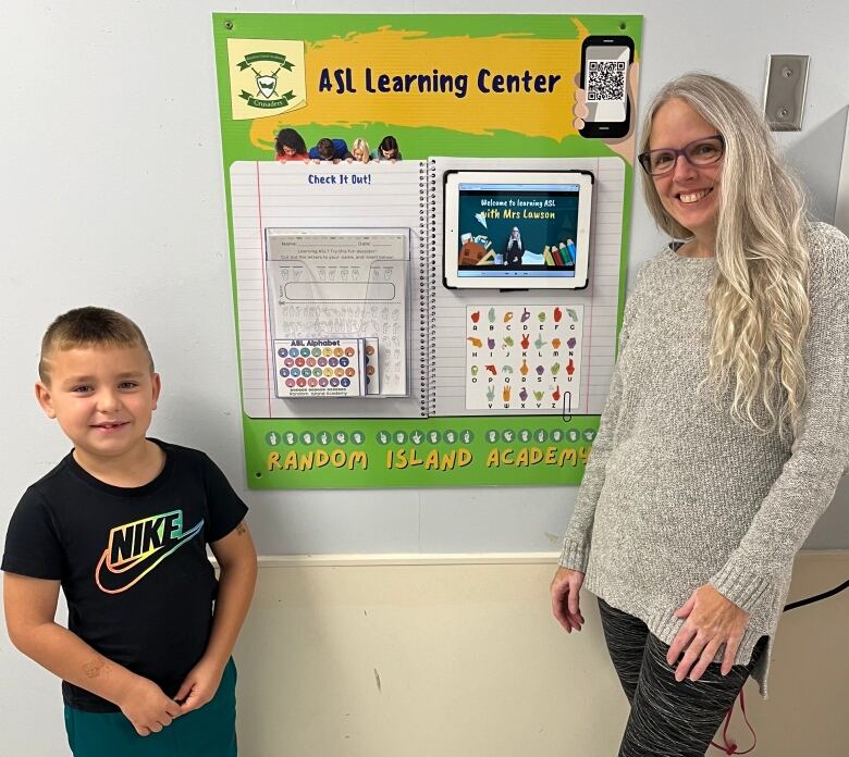 A young boy is standing next to a woman wearing glasses. Between them is a cardboard that says 