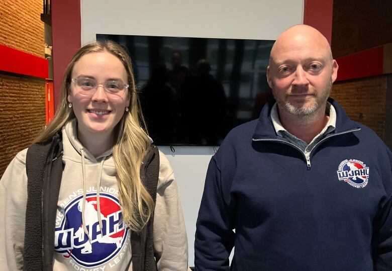 A young girl with long hair is smiling. She stands next to a bald man.