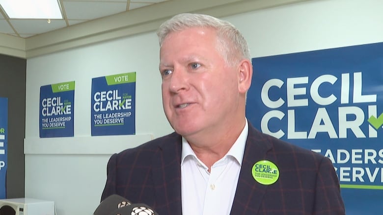 A man with silver hair, navy blue suit and white shirt wears a green button with the name Cecil Clarke on it in front of blue-green-and-white signs with the same name.