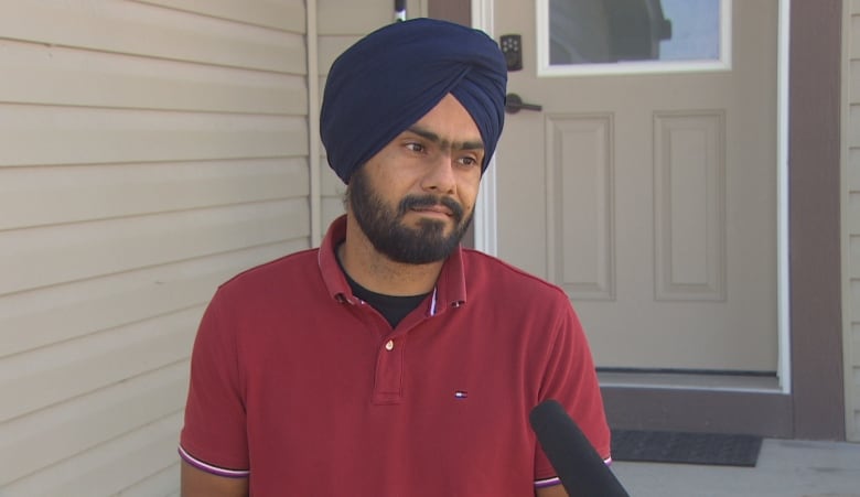 A man wearing a turban and a red shirt is pictured during an interview outside a home.