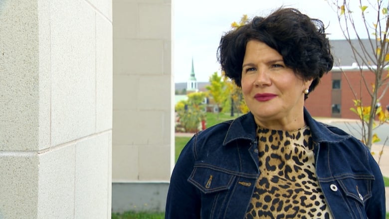 A white woman with a black bob and red lipstick wears a leopard-print shirt under a jean jacket. She stands outside with university brick buildings and trees behind her