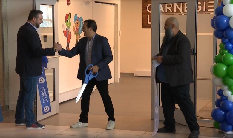 Two men shake hands in the doorway of a corporate office space, with a large pair of ribbon-cutting scissors.