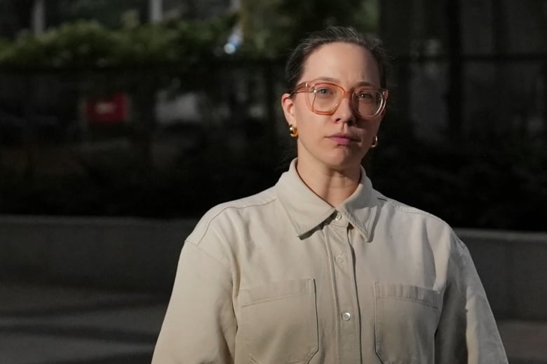 A woman in glasses and a light top poses for a photo against a dark background. 