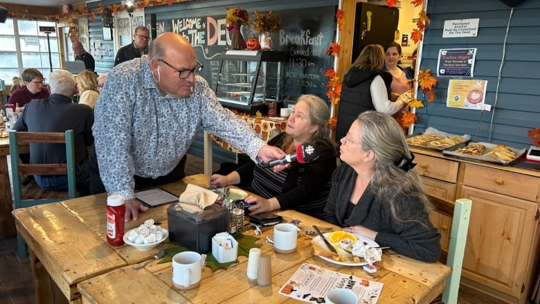 Man in a blue and white patterned shirt holds a microphone up to two women sitting at a table in a restaurant. 