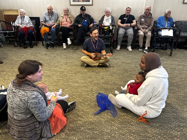 A group of people sits on carpet and on chairs.