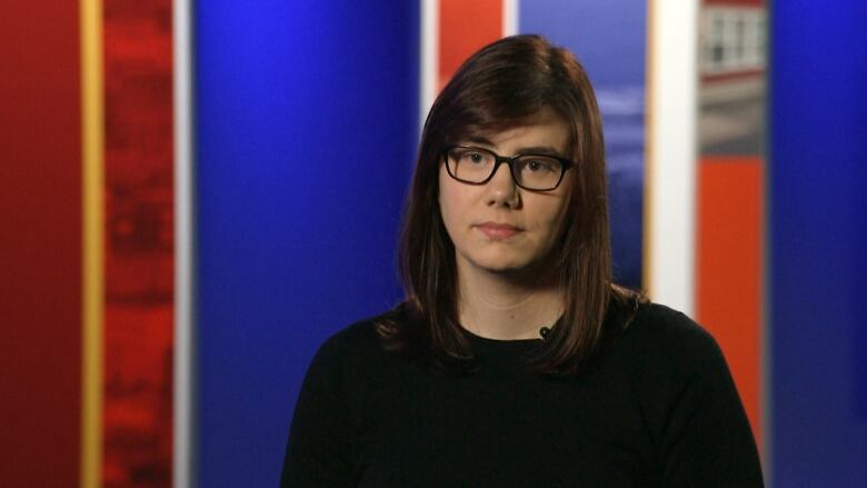 A woman with short brown hair and glasses stands in a news studio.