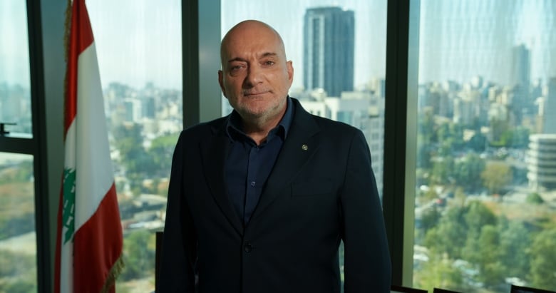 A man in a dark suit leans against a desk, in front of a Lebanese flag and a wide cityscape.