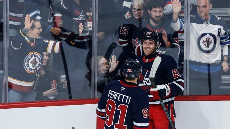 Two hockey players smile in celebration while fans behind the glass also celebrate.