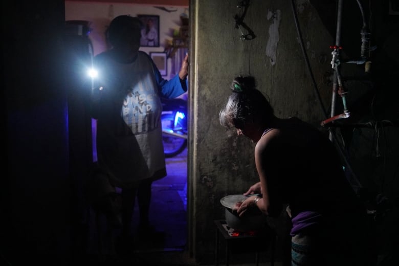 A person cooks on a stove amid a blackout as another person standing in the doorway uses a mobile phone's flashlight to illuminate the kitchen.