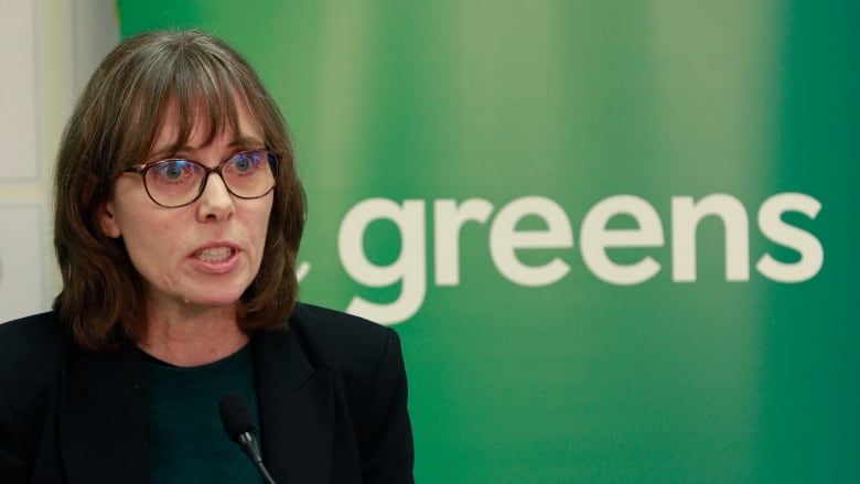 A woman speaks in front of a screen that reads 'Greens'.