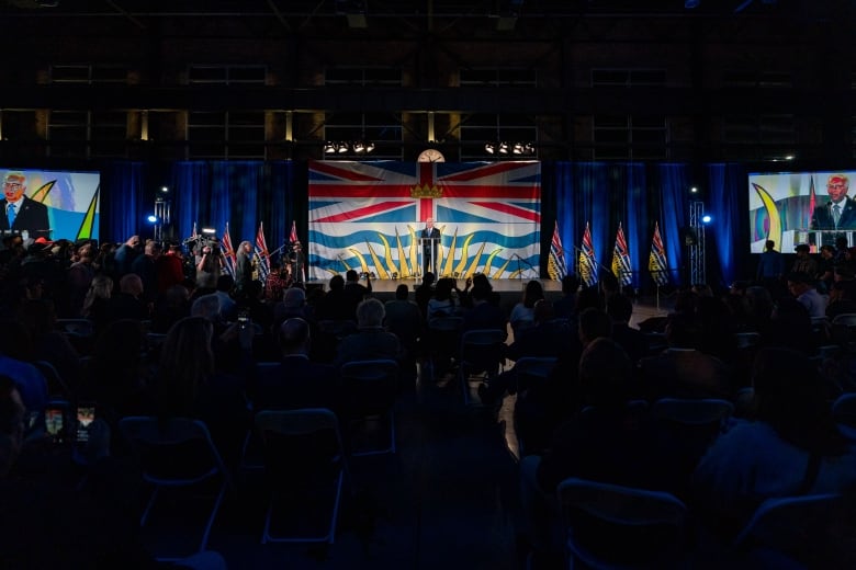 Dozens of people look at man in a suit on stage speaking behind a podium.