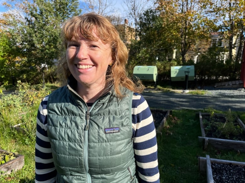 Woman with shoulder length blonde hair smiling, wearing a green vest and stripped shirt. Behind her is a garden with two green containers in the back next to some trees.