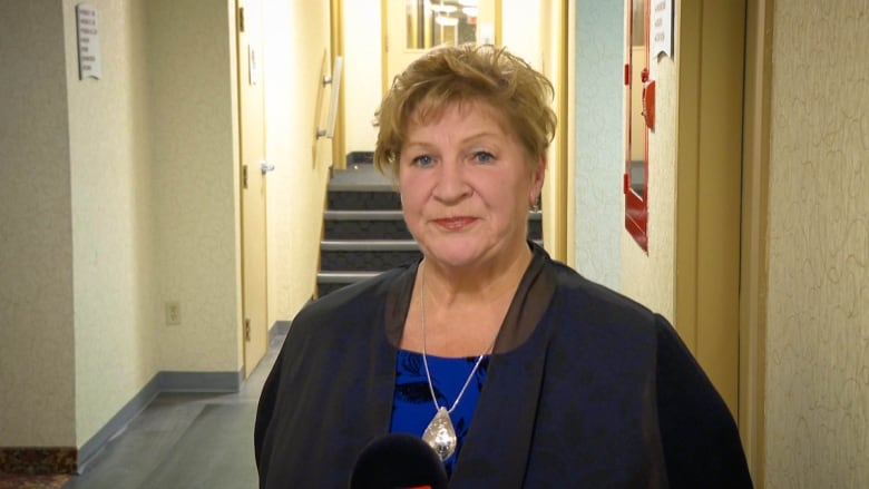 A woman with short brown hair, dark suit and blue shirt looks into the camera