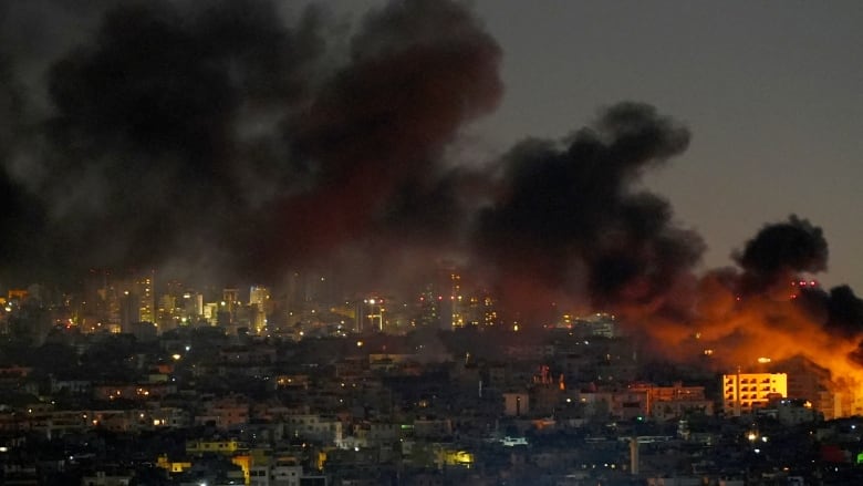 Black smoke stretches across the night sky above a city, rising from a building on fire in the far right of the image. 