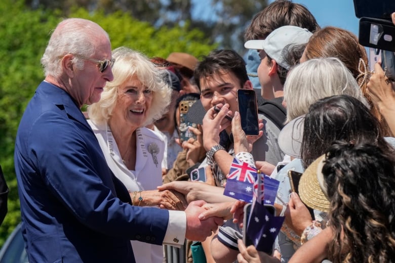 A smiling, elderly couple shakes hands with crowds of people who are lined up in a row. 