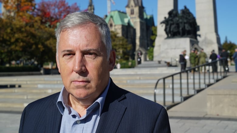 A man with white hair, wearing a navy jacket stands in front of the National War Memorial in Ottawa.