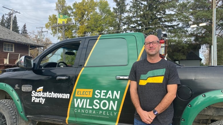 A man in a black shirt with a Saskatchewan flag logo stands in front of a black truck wiht a Saskatcehwan Party logo.