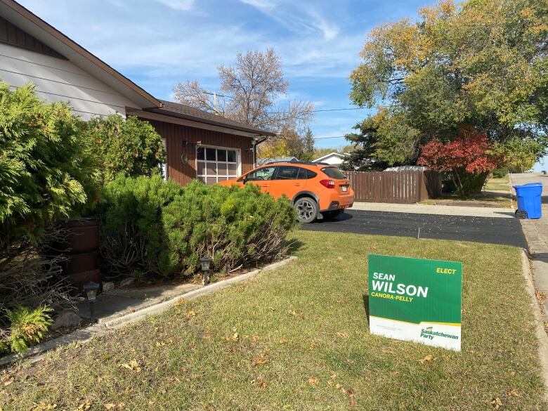 A green, white and yellow sign sits on a lawn with an orange car sitting on the driveway of the house.