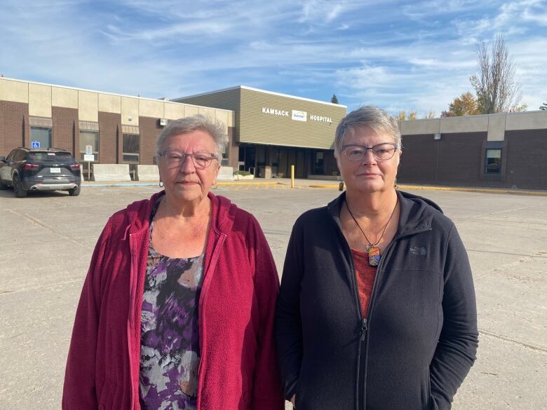 Two women with short white hair stand outside of a brown building that reads 'Kamsack hospital.' 