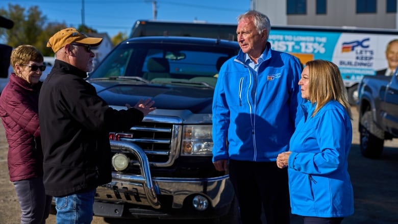 A man in a blue jacket beside a woman in a blue jacket standing in front of a pickup truck with two others on the left side of the frame.