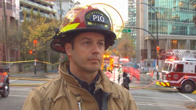 A firefighter speaks in a street with fire trucks and paraphernalia behind him.