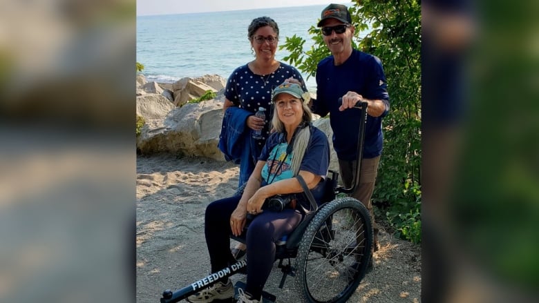 A woman sits in a wheelchair, with two people smiling behind her.