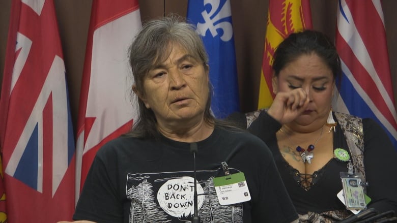 An Indigenous woman speaks as another woman behind her wipes away tears.