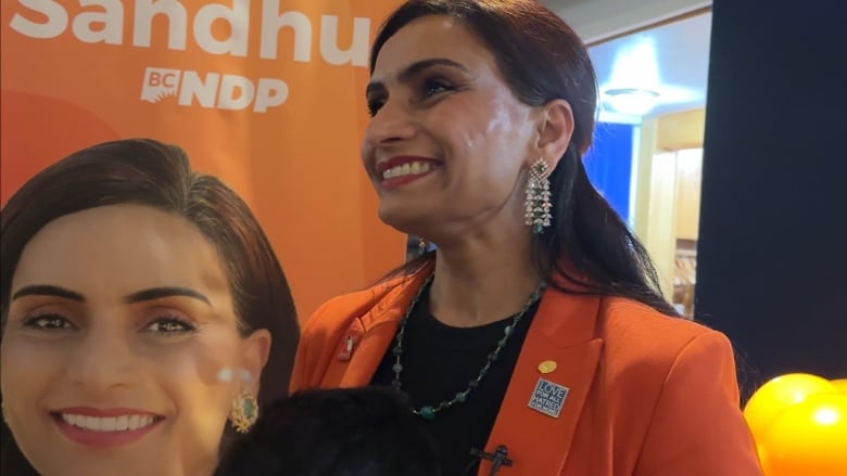 A smiling in an orange suit jacket stands in front of an NDP election sign.