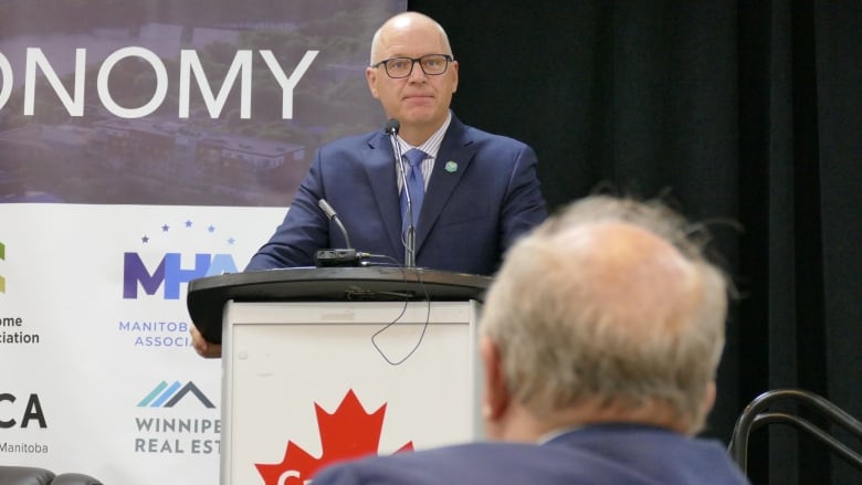 A bald man with glasses and a suit stands on a stage at a podium.