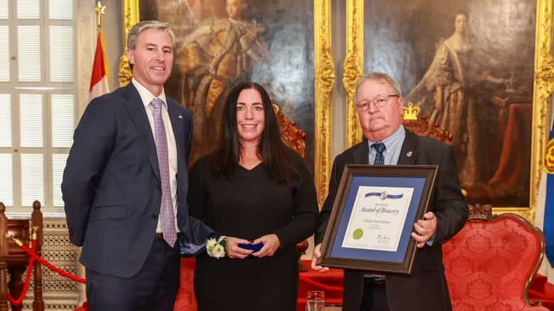 A woman holds a medal, to her left is a man in a suit and to her right is a man in a suit holding a framed certificate. 