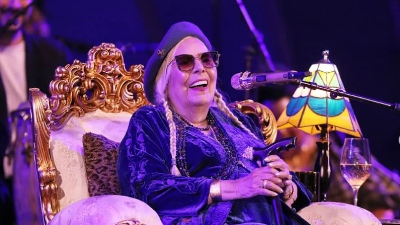 Joni sits on her throne while wearing a dark velvet dress and hat.