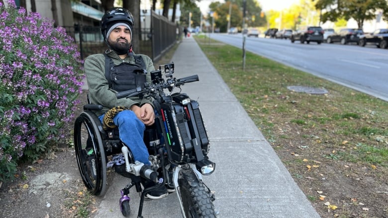 Man sitting in a wheelchair on the sidewalk beside a road.