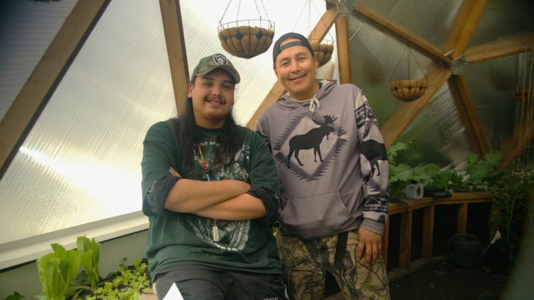 Two Indigenous youth smile at the camera, surrounded by plans in a geodesic dome greenhouse.