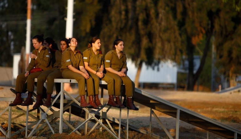 Roughly six women, in military uniforms, sit outdoors on a large ramp. 