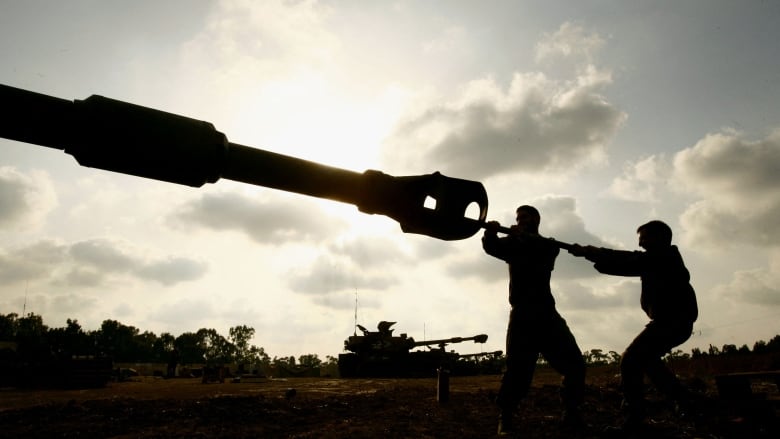 Two men, in silhouette, push a large pole into the main gun of a tank. 