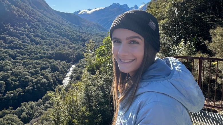 A woman wearing a beanie smiles for a photo in front of a mountain range. 