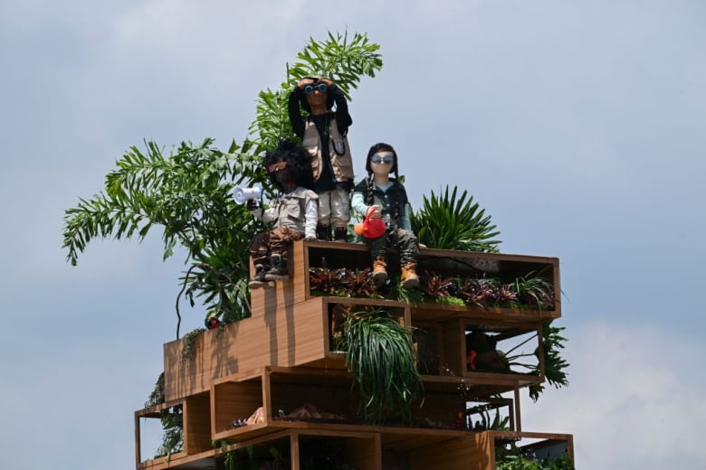 Sculpture of palm fronds and three children sitting on a platform made of offset hollow rectangular wood blocks. The children have no mouths. One is peering through binoculars. One has a megaphone held up to its face. And one is holding a watering can.