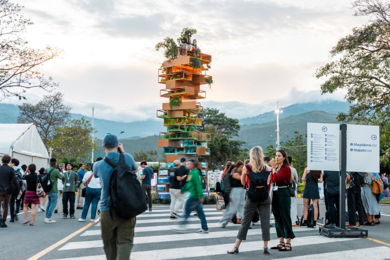 People stand and walk in the streets in front of a tall sculpture, shaped like a Jenga tower and made of hollow wooden boxes. Plant life spills out of the boxes, and sculptures of people sit atop it.