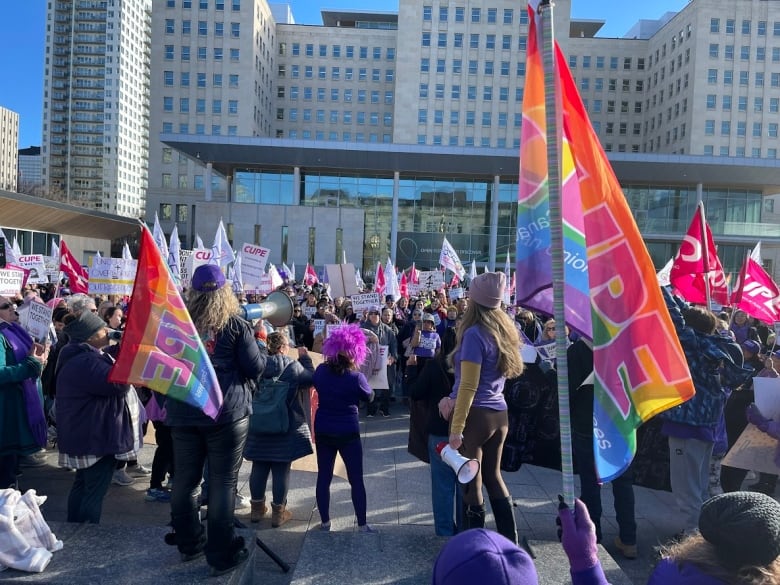 A group of people rallying, some with CUPE rainbow flags, with one person in the middle holding a megaphone. 