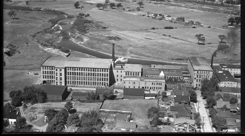 A black and white aerial photo shows a series of factory buildings on a riverbank, with smoke coming from a tall chimney