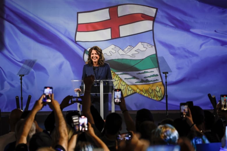 A woman stands in front of an Alberta flag in front of a crowd.