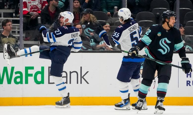 Hockey players in white and blue celebrate a goal while an opposing player in teal and navy skates past.