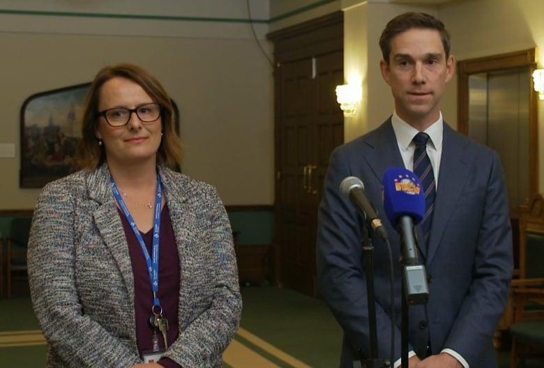 A man and woman stand outside the House of Assembly in front of two microphones.