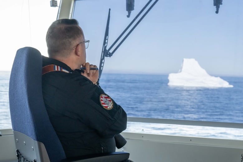 A military ship captain looks out of the ship's window at an iceberg nearby.