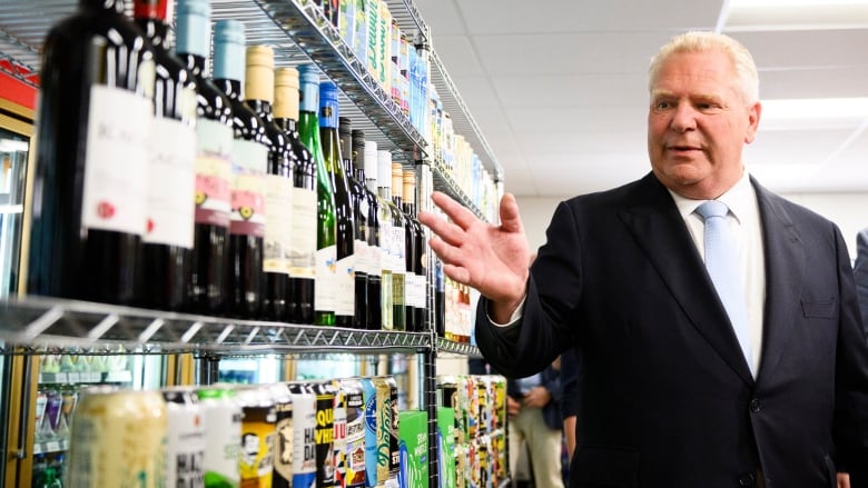 A man in a suit stands at the right side of the image, gesturing towards a shelf full of alcoholic beverages on the left side of the image. There is an entire shelf of just wine bottles, and then other beverages are visible in the shelves below and above. 