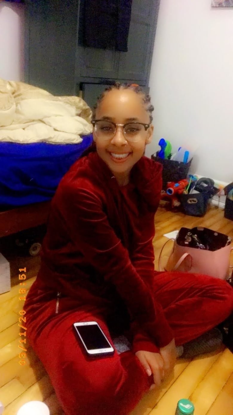 A young woman sitting on a floor and smiling for the camera. She is wearing a red jumpsuit and glasses.