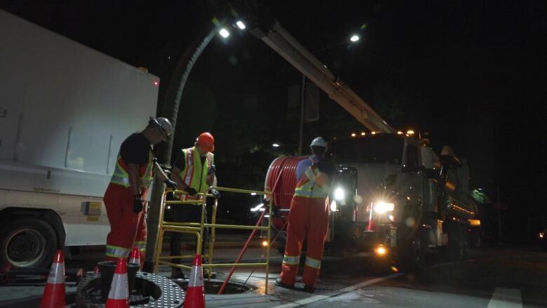 Three workers in reflective suits and gear work with a truck in the background surround a manhole with traffic cones to bar access to public.